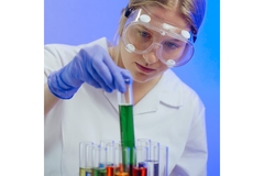 Image A woman wearing protective goggles and a lab coat picking up a test tube filled with colorful liquid.